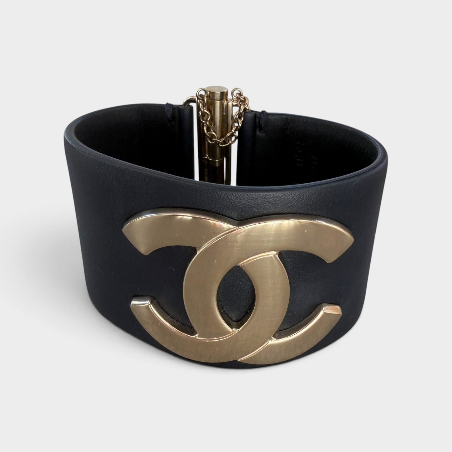 A wide black leather cuff bracelet featuring a large gold-tone interlocking CC logo on the front, with a gold-tone chain closure visible at the top, photographed on a light gray background.