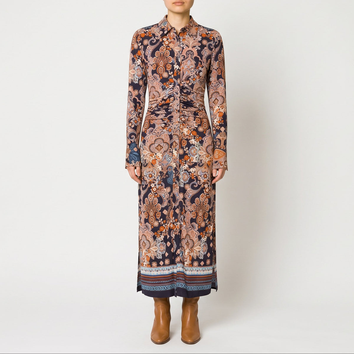 A model stands against a light gray studio background wearing a long-sleeved midi shirt dress with an intricate multicolor paisley and floral pattern in shades of brown, orange, beige, and navy, paired with brown ankle boots.