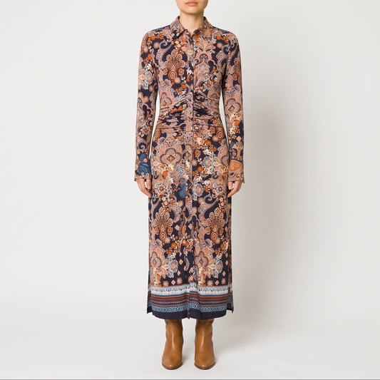 A model stands against a light gray studio background wearing a long-sleeved midi shirt dress with an intricate multicolor paisley and floral pattern in shades of brown, orange, beige, and navy, paired with brown ankle boots.