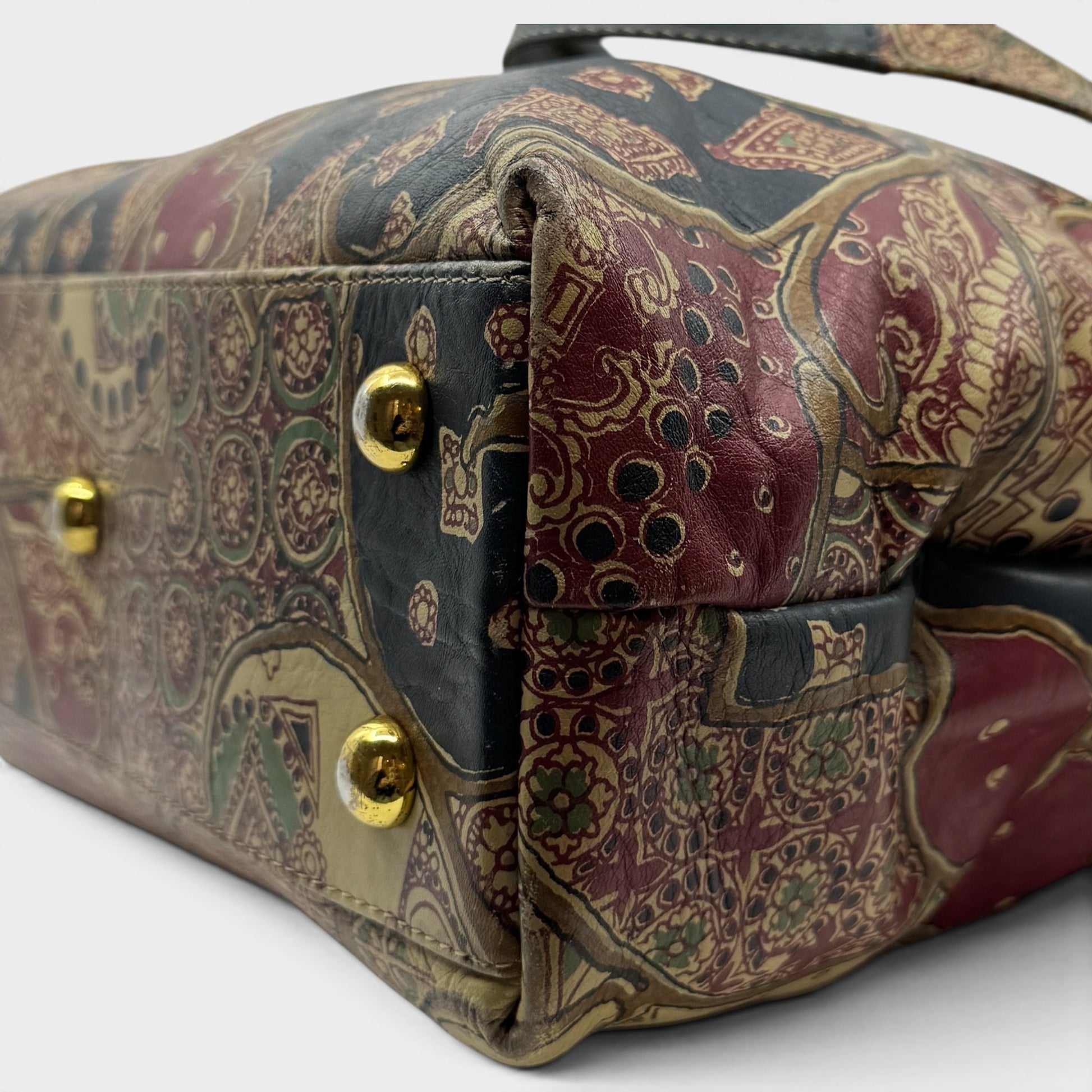 A close-up of a handbag corner showing a detailed paisley pattern in burgundy, gold, and black, with gold-tone metal studs.